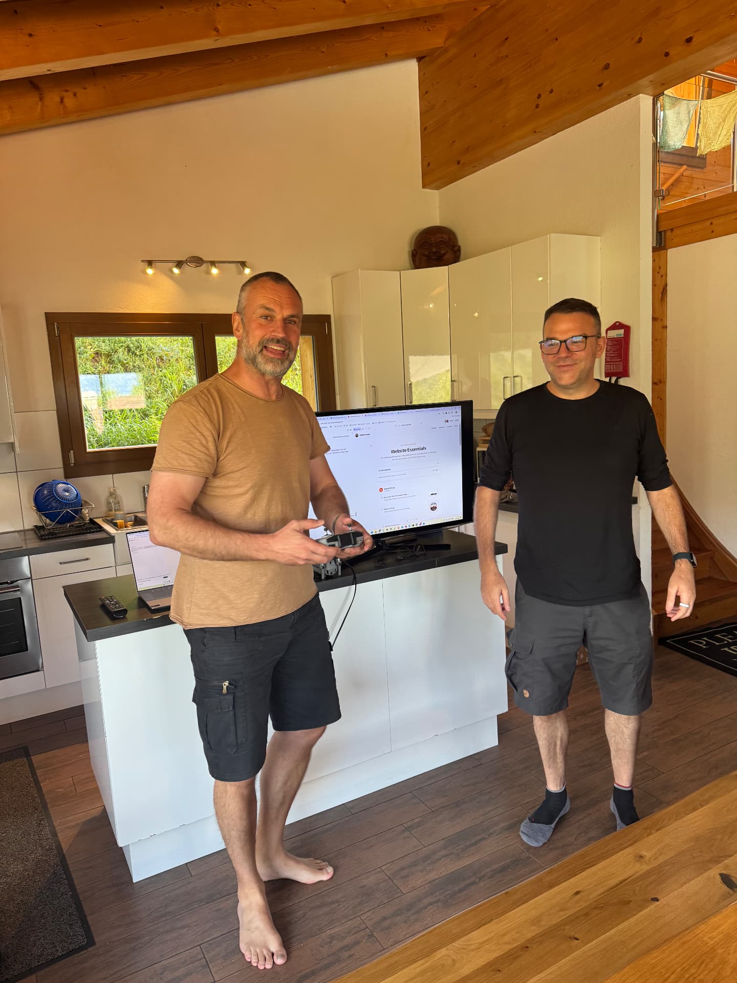 The two VibeMountain hosts in the chalet kitchen next to a screen showing the website